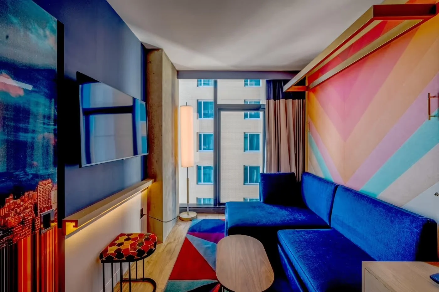 A modern hotel room with a large window, a blue velvet sofa, colorful geometric-patterned wall art, a floor lamp, a flat-screen TV, and contemporary furniture.