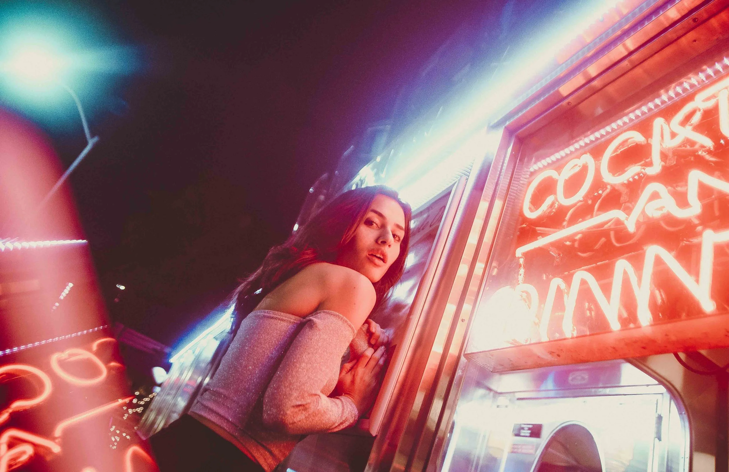 A young woman with long hair, wearing a long-sleeve top, posing in front of red neon lights at night.