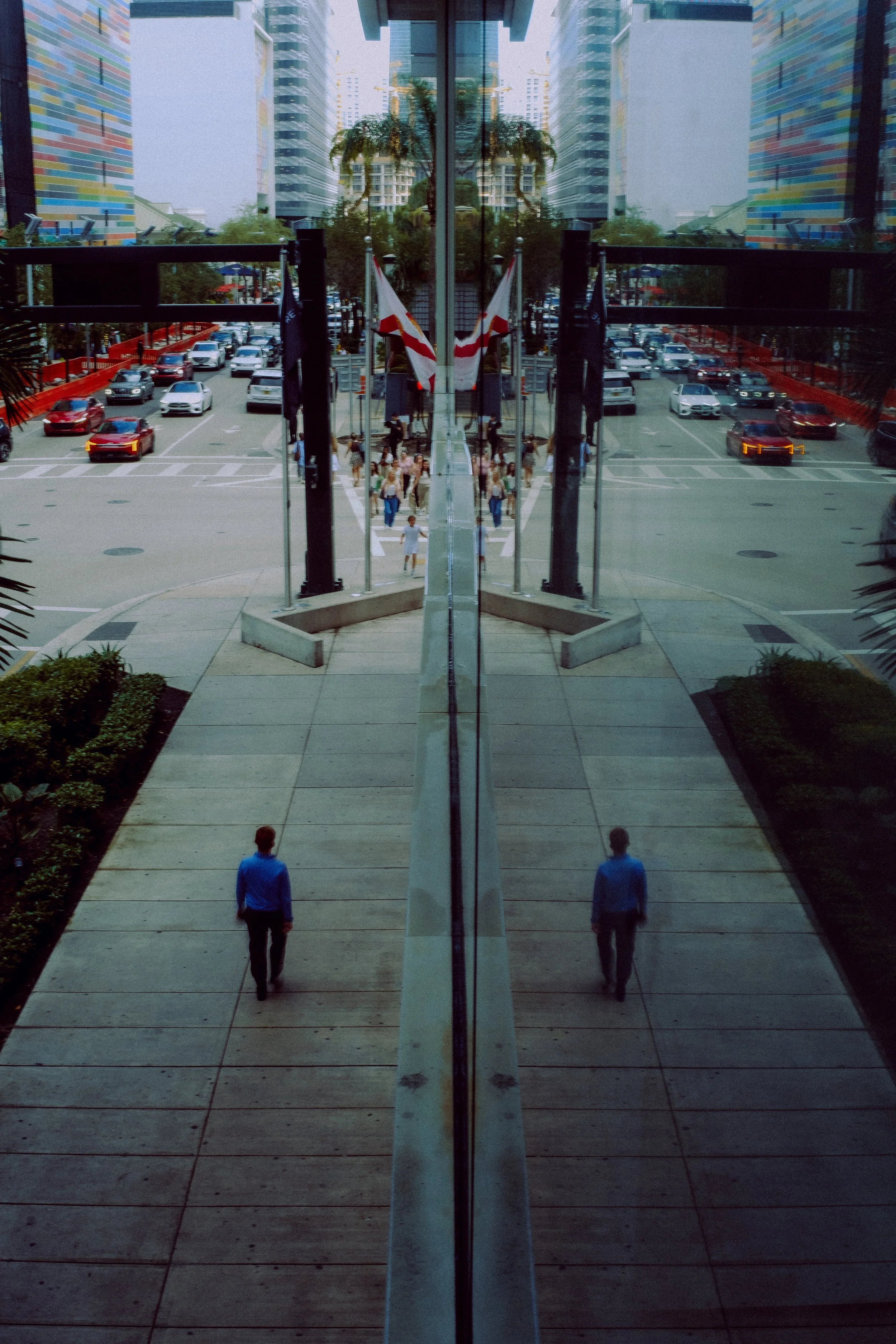Reflected view of a city street through glass doors, with people walking on the sidewalk and cars parked along the street. The reflection creates a mirror effect with buildings and a palm tree visible in the glass.
