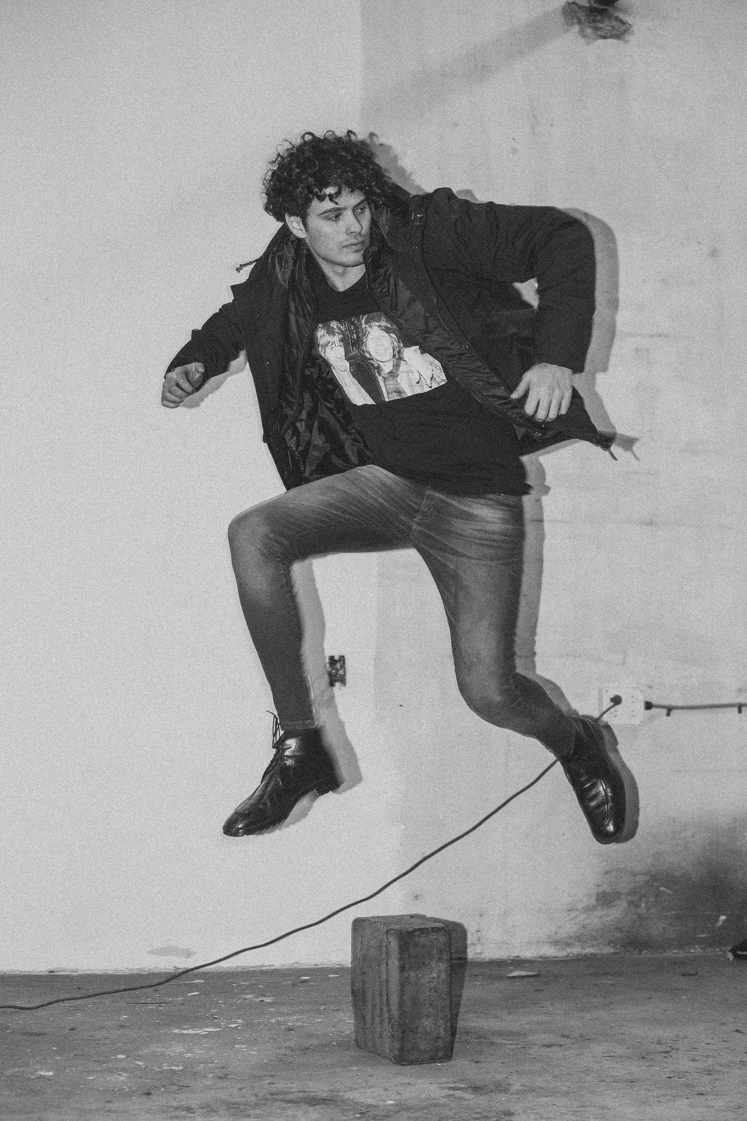A person with curly hair wearing a jacket, graphic T-shirt, jeans, and dress shoes, posed in front of a plain wall. They are slightly leaning to the side with one leg raised, with a cable running across the floor attached to a small speaker.