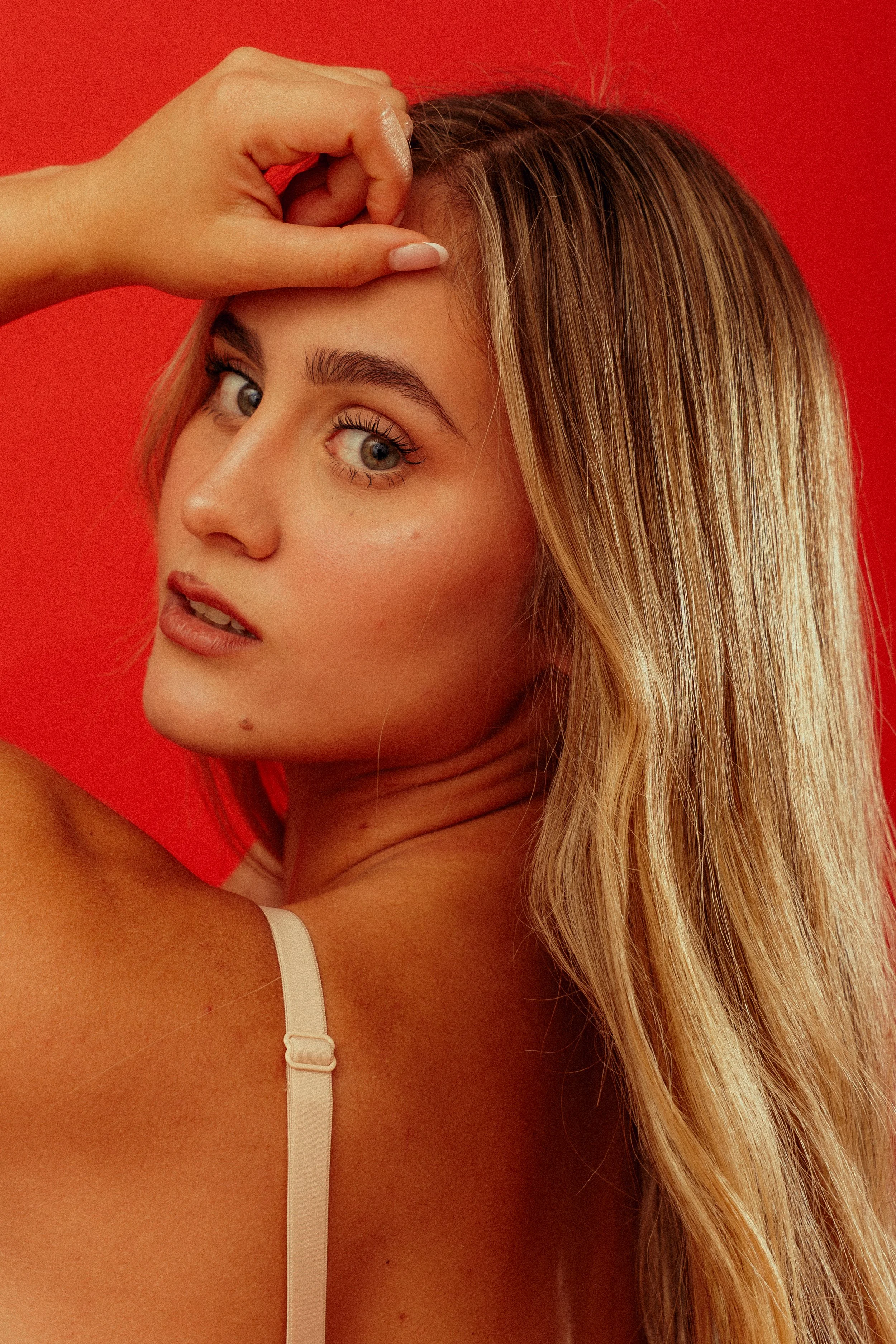 A woman with long blonde hair looking at the camera, with her hand raised near her forehead, against a red background.
