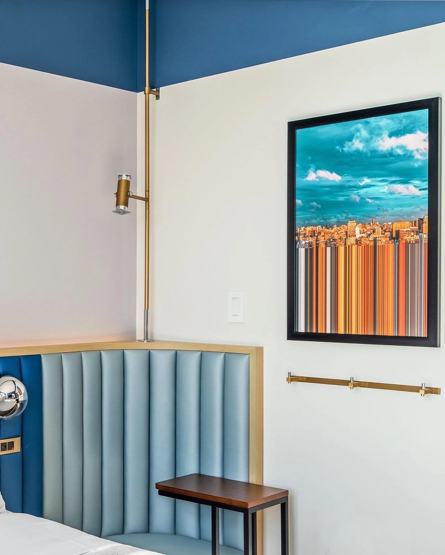 Interior of a modern bedroom with a blue and white color scheme, featuring a patterned headboard, a framed cityscape photograph with a cloudy sky, modern wall-mounted lighting, and a brass railing.