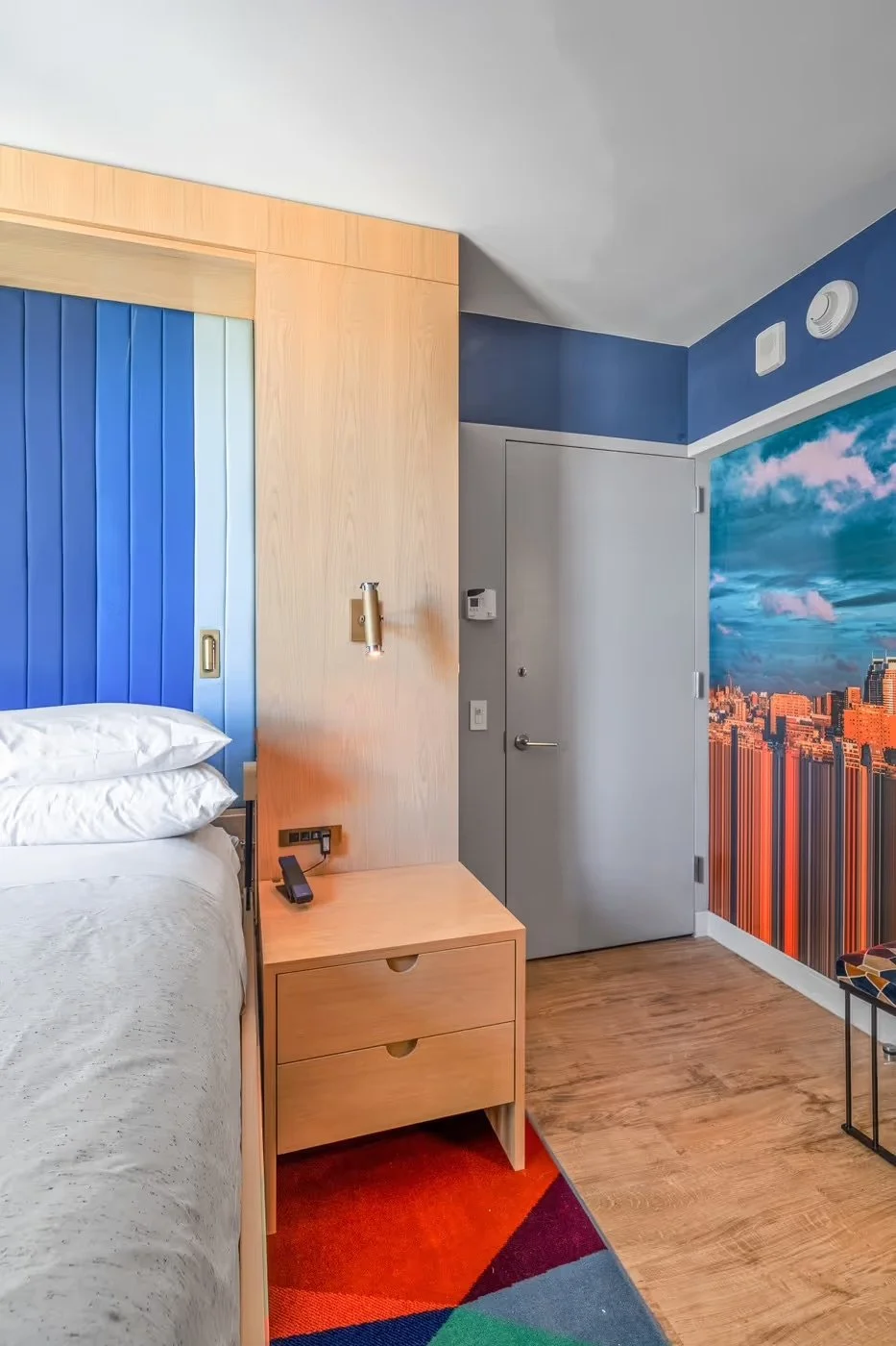 Part of a hotel room showing a bed with white linens, a bedside table with a phone, a wall-mounted reading light, a door, a mural of a city skyline under a cloudy sky, and a colorful geometric rug.