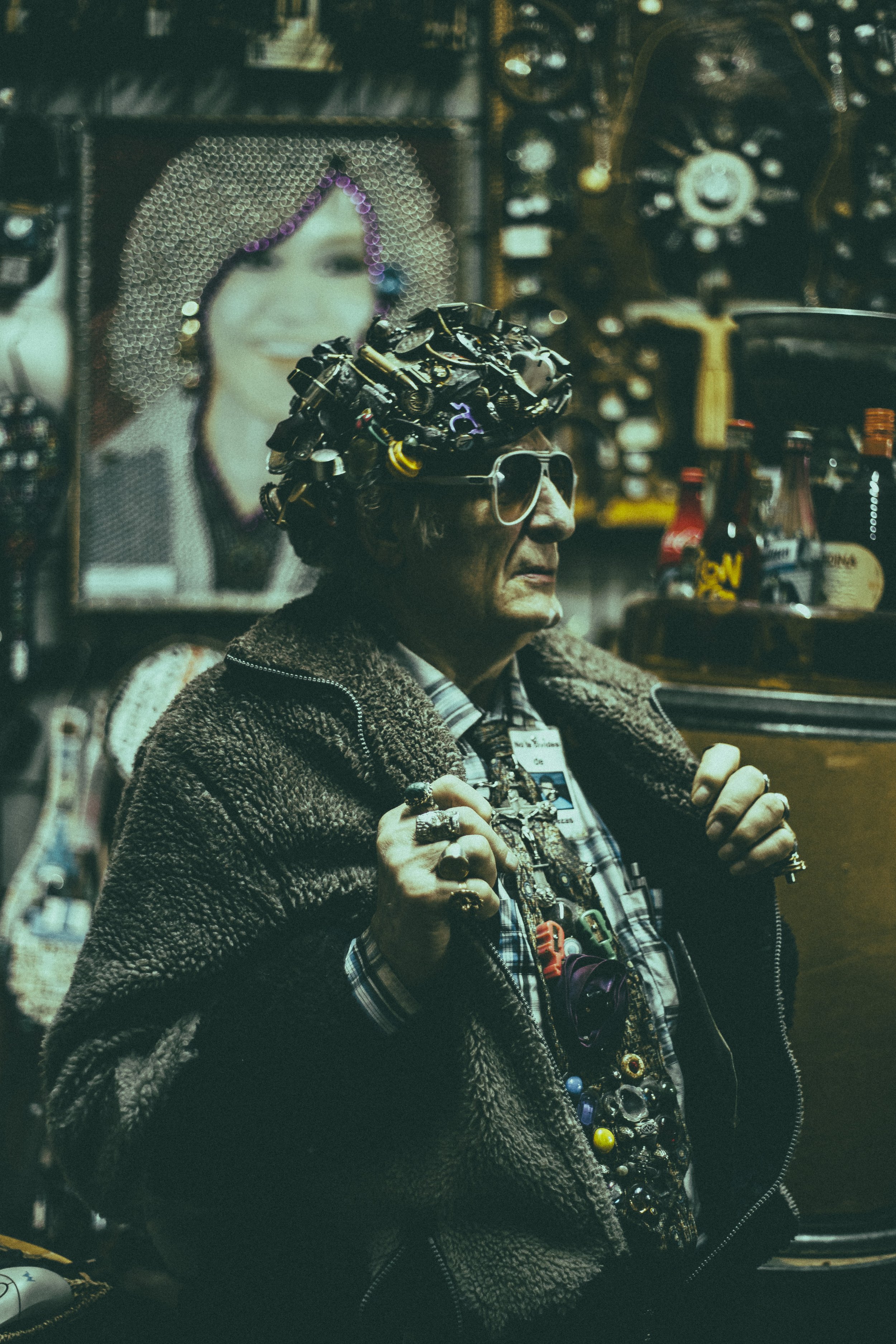 A person wearing sunglasses, a plaid shirt, and a jacket with a fuzzy collar, sitting in front of shelves filled with various bottles and objects, and a large framed photo or poster of a smiling woman with curly hair in the background.