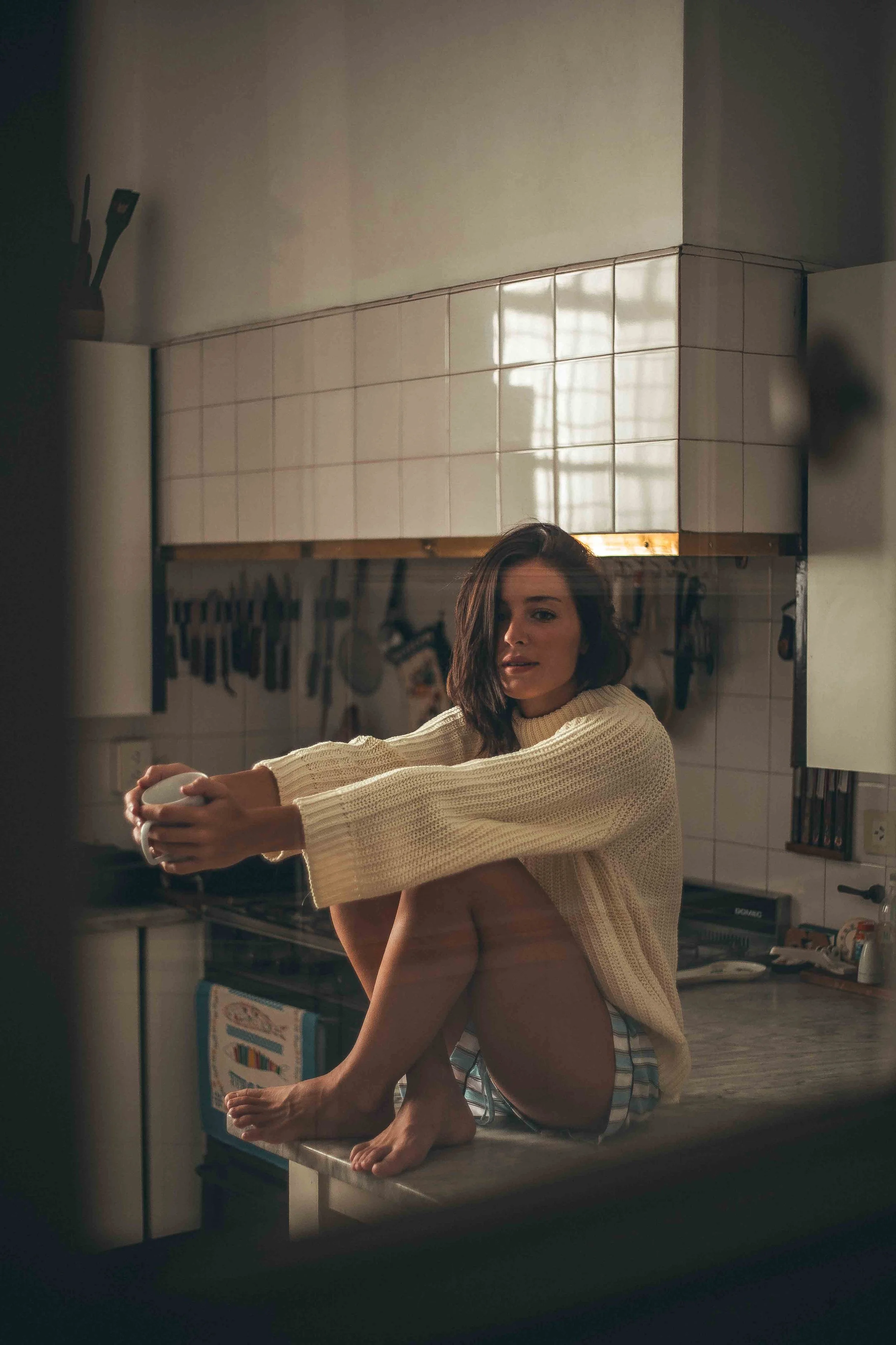 A woman with brown hair and a striped shirt sitting on a kitchen countertop holding a mug, with her knee raised. The kitchen has white tiled walls and hanging utensils in the background, with warm lighting and a reflection of a window on the wall.