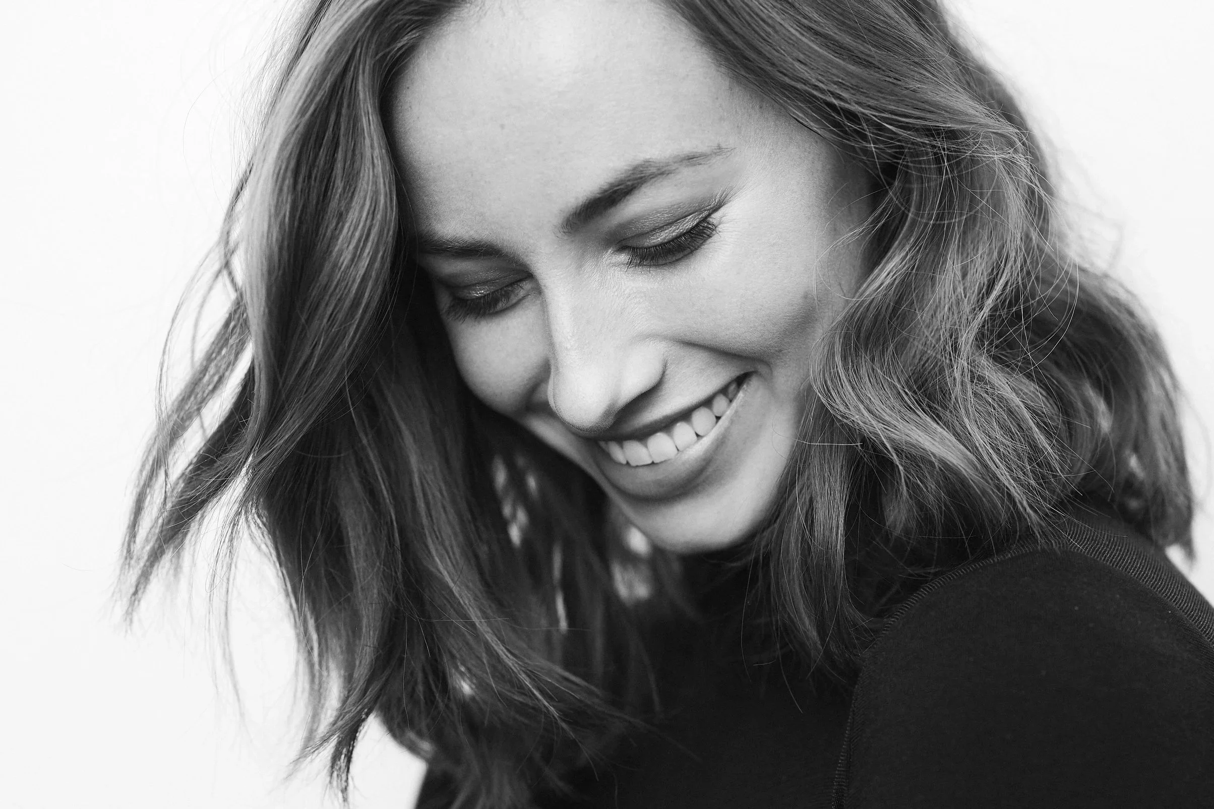 Black and white close-up portrait of a young woman smiling with eyes closed, long wavy hair, and clear skin.