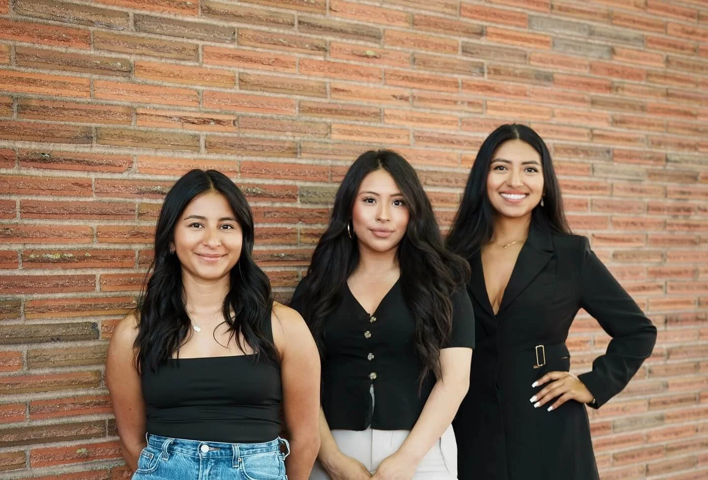 Three women standing side by side against a brick wall, smiling at the camera.