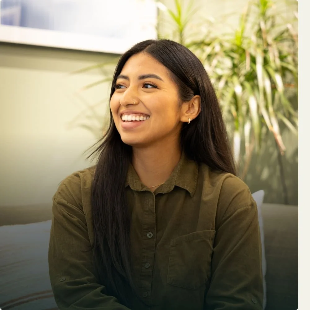 A young woman with long dark hair smiling and looking to her left, wearing a green button-up shirt, seated indoors with a blurred background of green plants and a window.