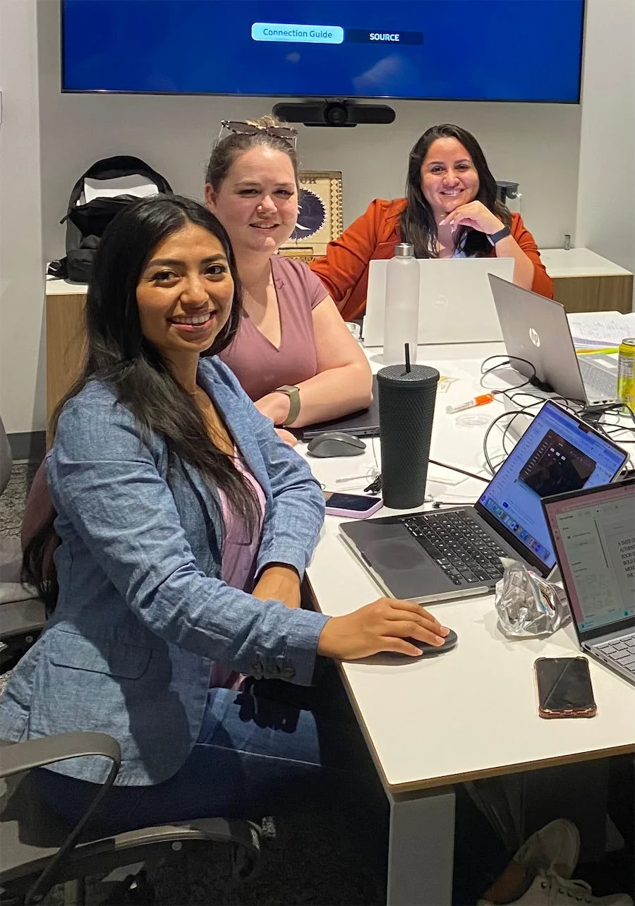 Three women sitting at a table in a meeting room with laptops, water bottles, and workspace items, smiling at the camera.
