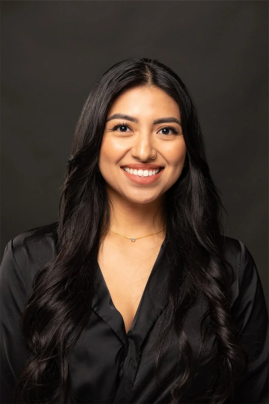 A young woman with long dark hair, smiling, wearing a black satin shirt and a delicate gold necklace with a small gemstone, against a dark background.