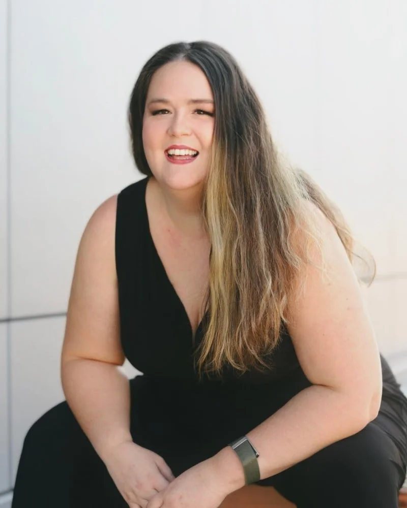 A woman with long, wavy hair, wearing a black sleeveless top, sitting indoors against a plain white wall, making a playful facial expression.