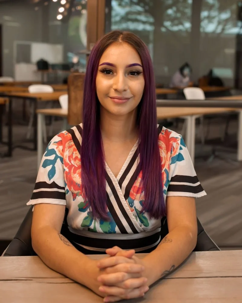 A young woman with long purple hair sitting at a table in a casual indoor setting, smiling softly at the camera.