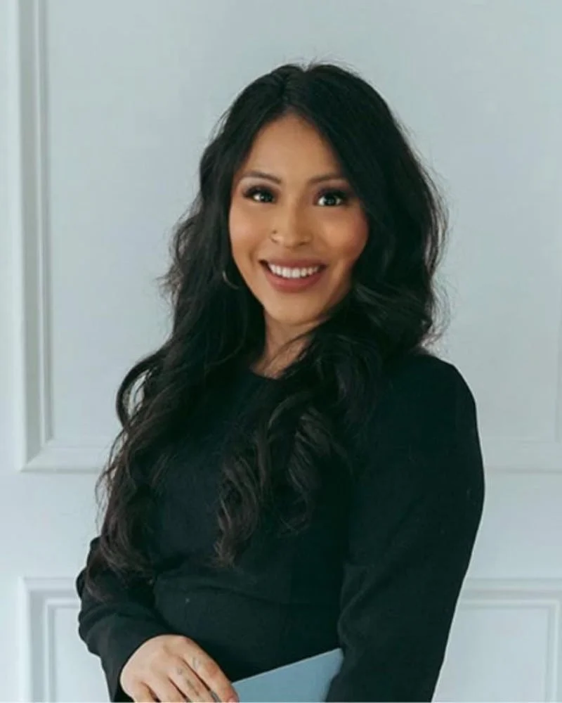 A woman with long black wavy hair, wearing a black top, smiling and holding a folder, standing in front of a white paneled wall.