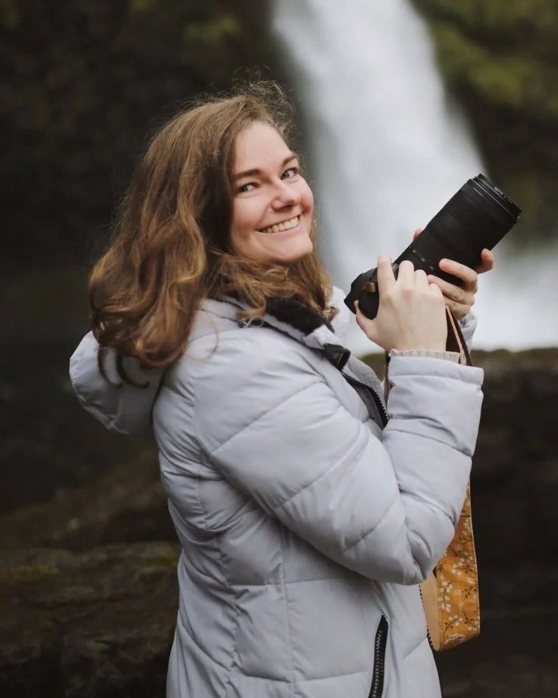 A woman with curly brown hair smiling and holding a camera outdoors, with a waterfall in the background.