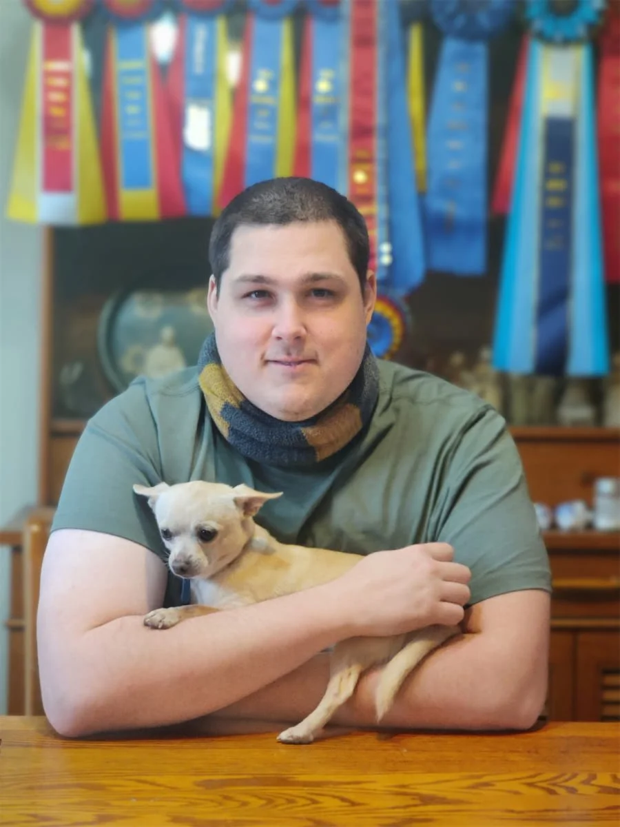 A man with short dark hair and a scarf around his neck holds a small tan and white dog in his arms, sitting at a wooden table inside a room decorated with colorful rosettes or ribbons hanging on a wall in the background.