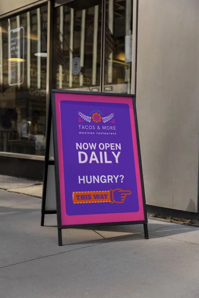 A sidewalk signboard outside a restaurant named "Tacos & More" announcing it is now open daily with a prompt asking if viewers are hungry, featuring an orange arrow and hand icon pointing to the direction.
