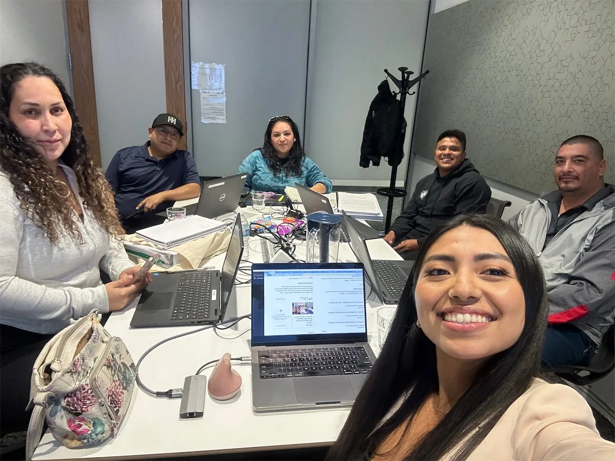 Group of six people sitting around a conference table with laptops, notebooks, and drinks, taking a selfie in a modern office meeting room.