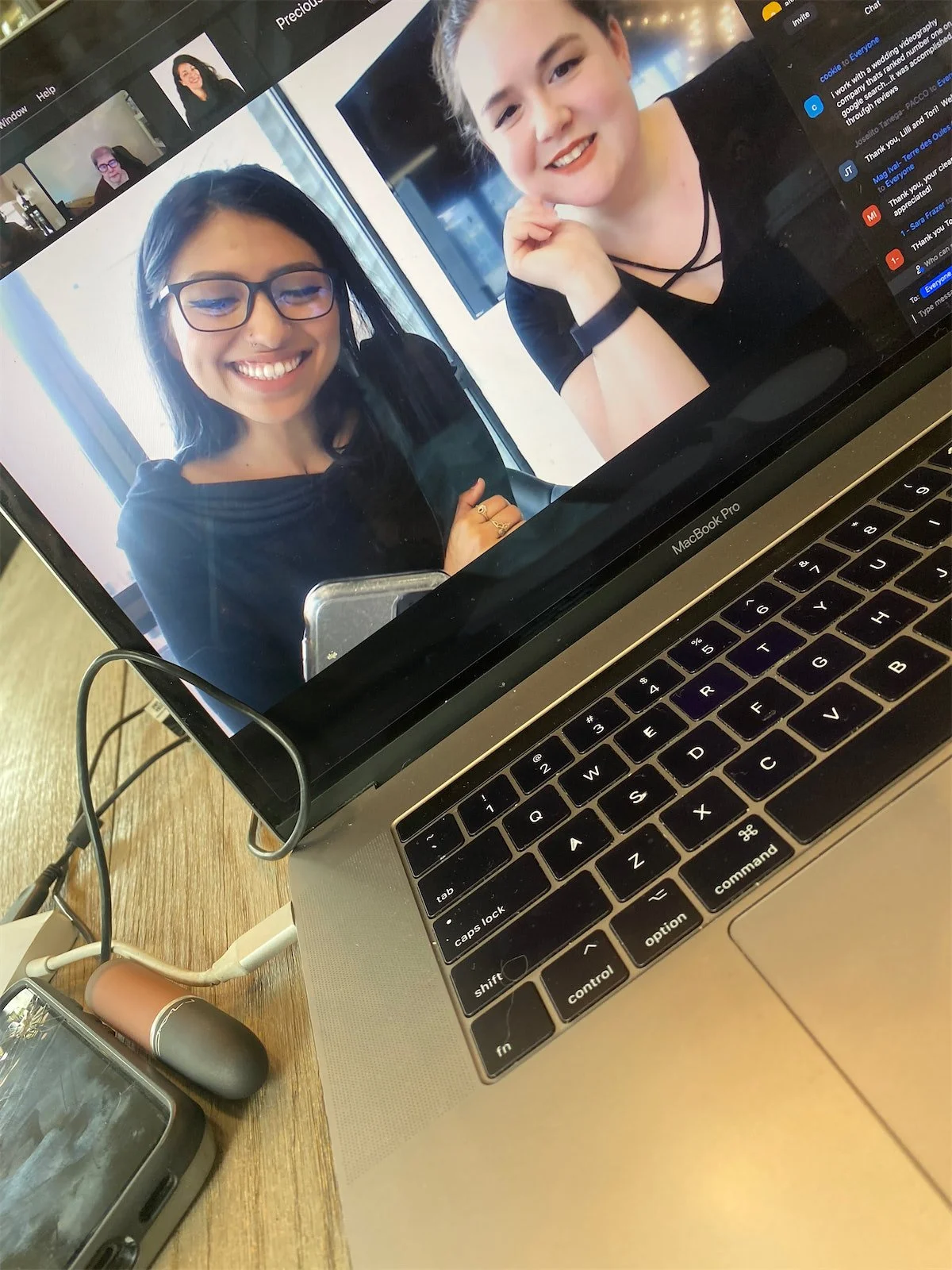 A laptop screen showing two women smiling during a video call, one woman has dark hair and glasses, and the other has light hair and is wearing a black top. The laptop is placed on a wooden table with phones and charging cables nearby.
