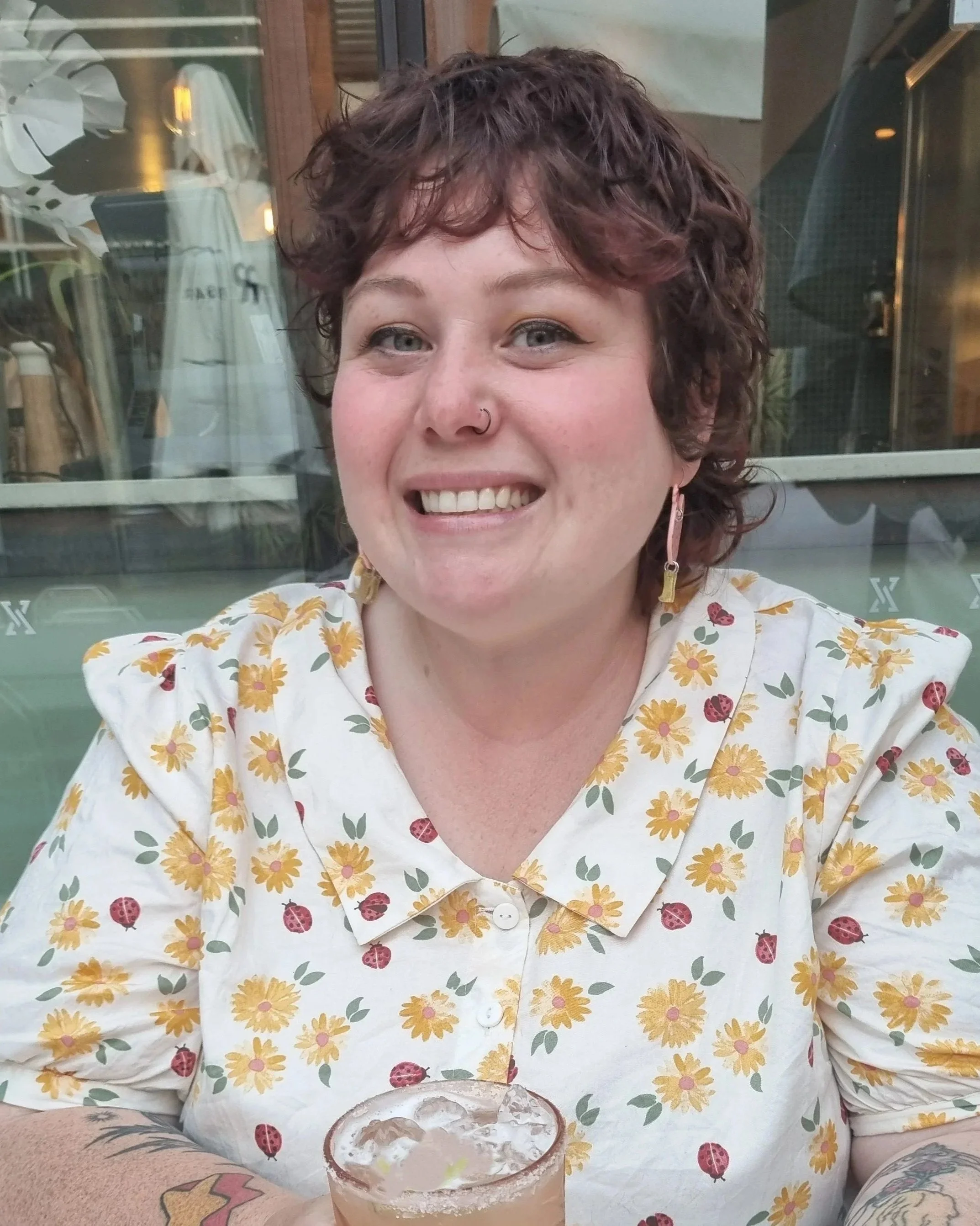 A smiling woman with short curly brown hair, wearing a floral blouse, sitting at an outdoor table with a cocktail drink that has a salted rim and lime garnish. Behind her is a window showing reflections and indoor items.