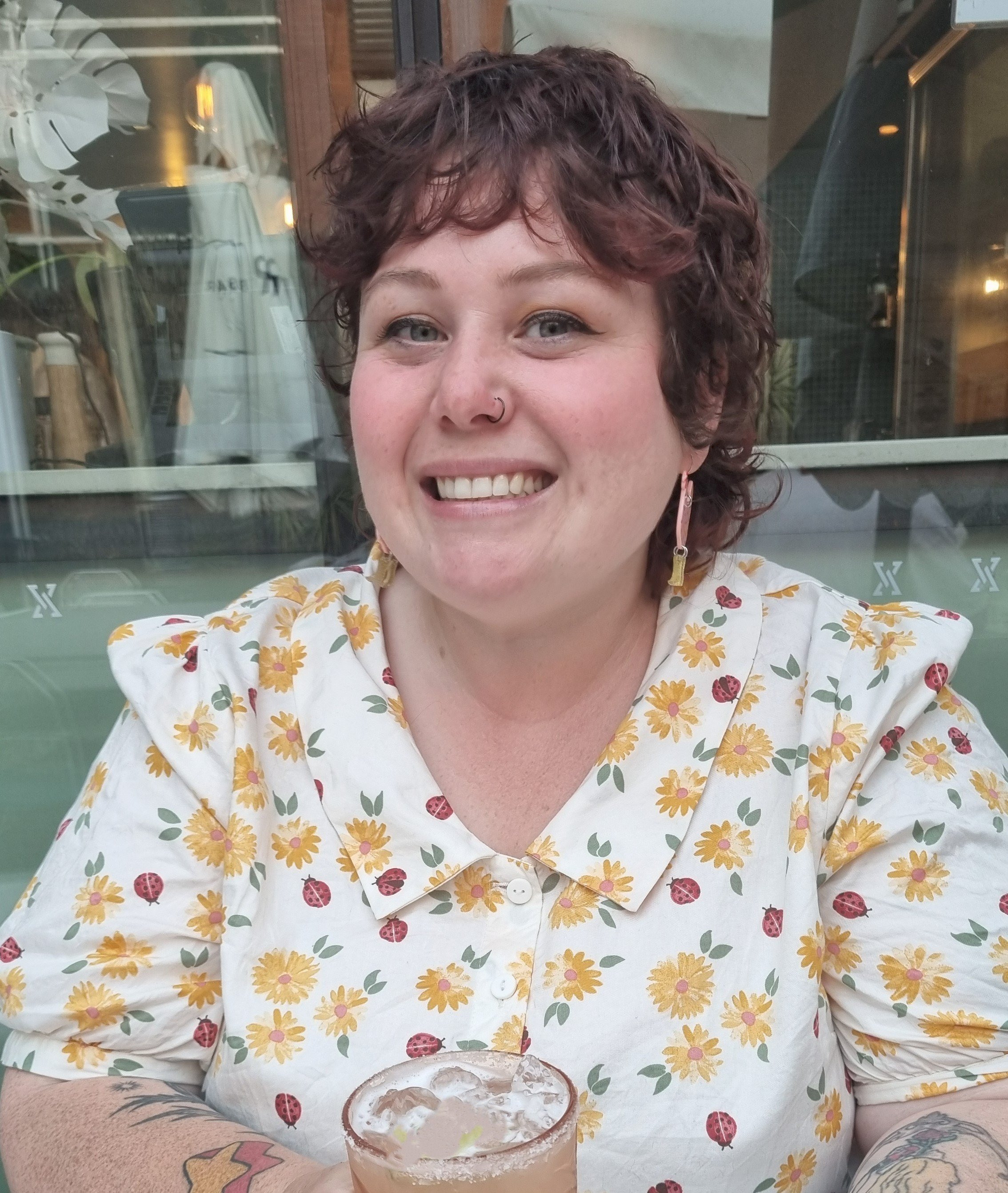 A woman with short, curly brown hair and nose ring smiling at the camera. She is wearing a white shirt with yellow and red floral and ladybug pattern. In front of her is a glass of a drink with a salted rim and lime slices. She has tattoos on her arms. The background shows a building with glass windows and reflections.