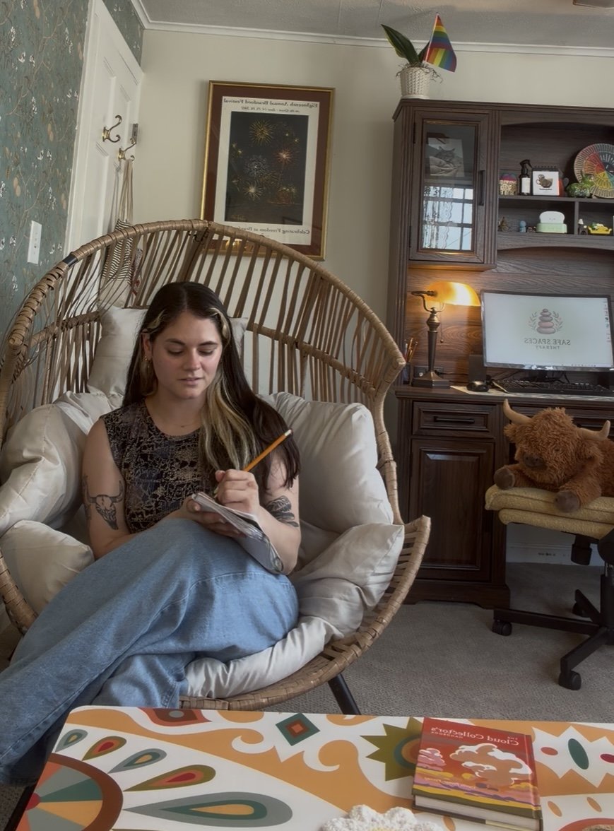 A young woman with tattoos on her arms sitting in a wicker chair in a cozy room, writing in a notebook with a pencil, surrounded by pillows. Behind her, there's a framed picture of fireworks, a wooden cabinet with decorative items, and a desk with a mouse and monitor displaying a spa logo. A plush teddy bear is on a chair next to the desk, and a rainbow party hat is on top of a white basket.