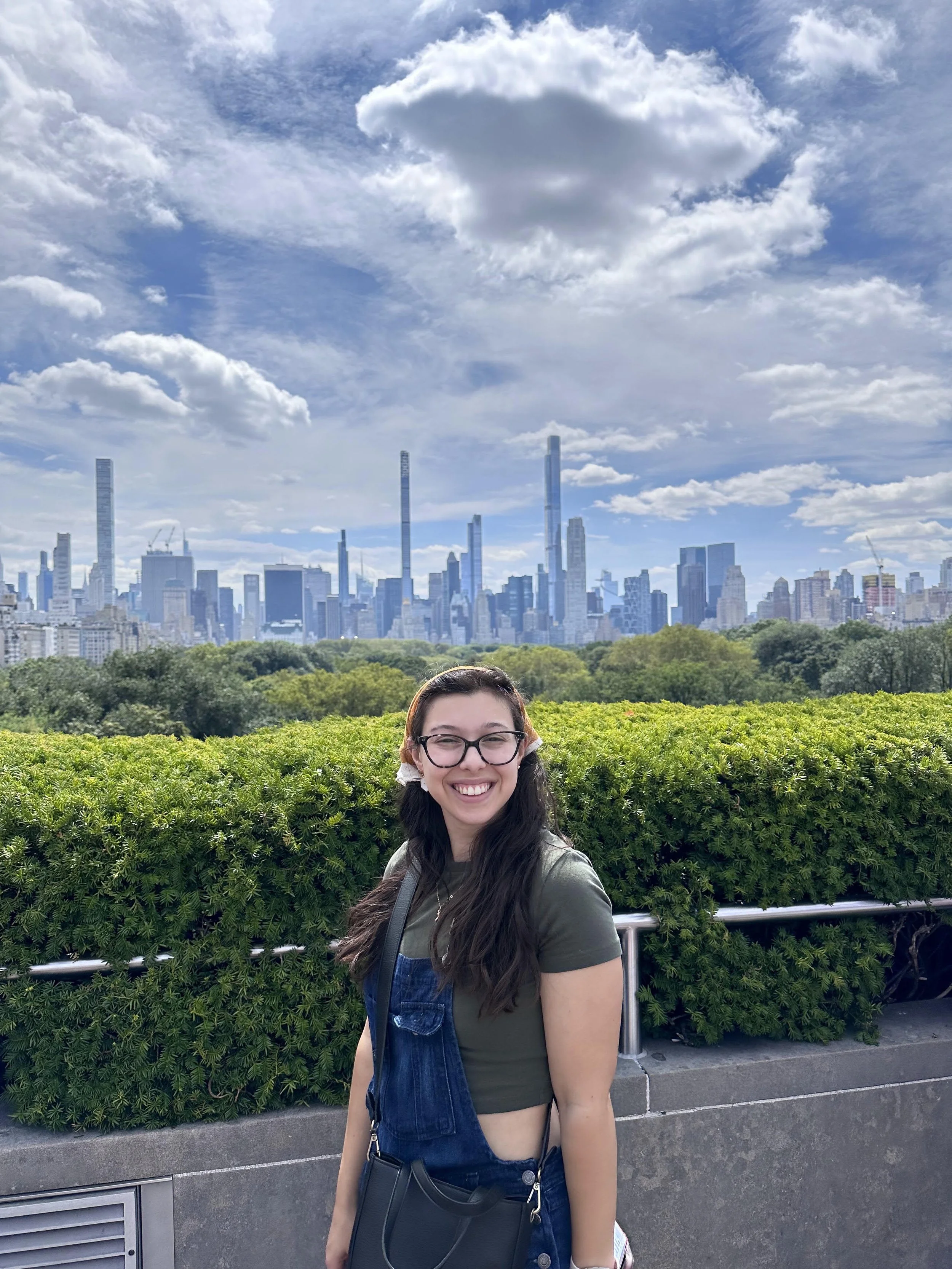A young woman smiling outdoors with the New York City skyline in the background, including skyscrapers and a partly cloudy sky.