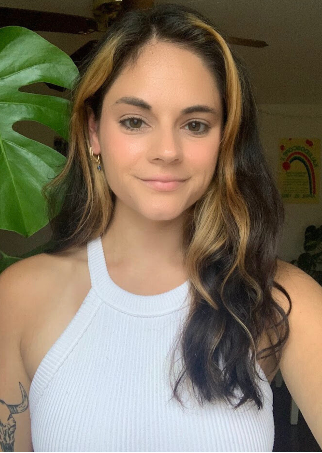 Close-up of a young woman with wavy, blonde and black hair in a white sleeveless top, standing in front of a green plant and a colorful rainbow poster.