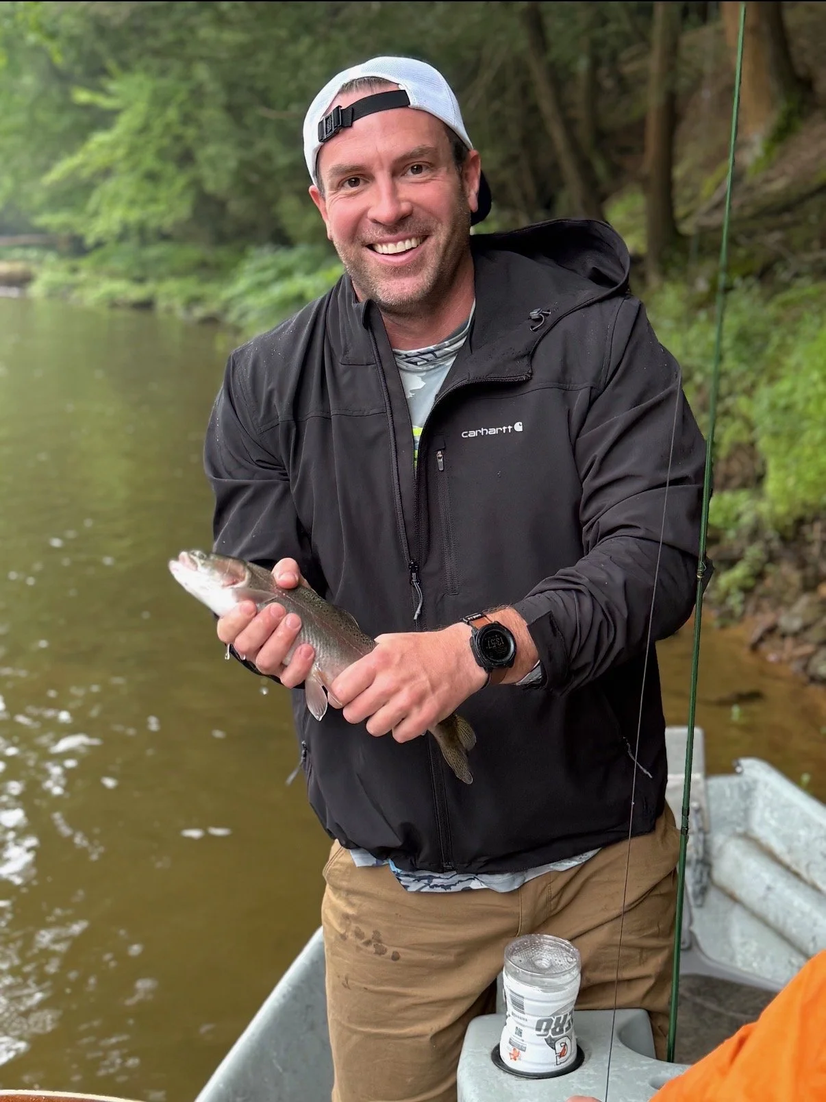 A man smiling while holding a fish he caught, standing in a boat by a river with green trees in the background.