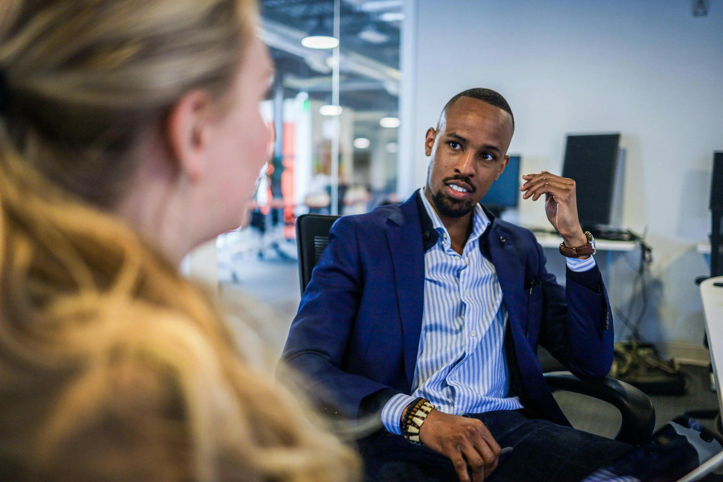 A man in a blue blazer, striped shirt, and jewelry is engaged in a conversation with a woman in an office environment with computers and glass walls.