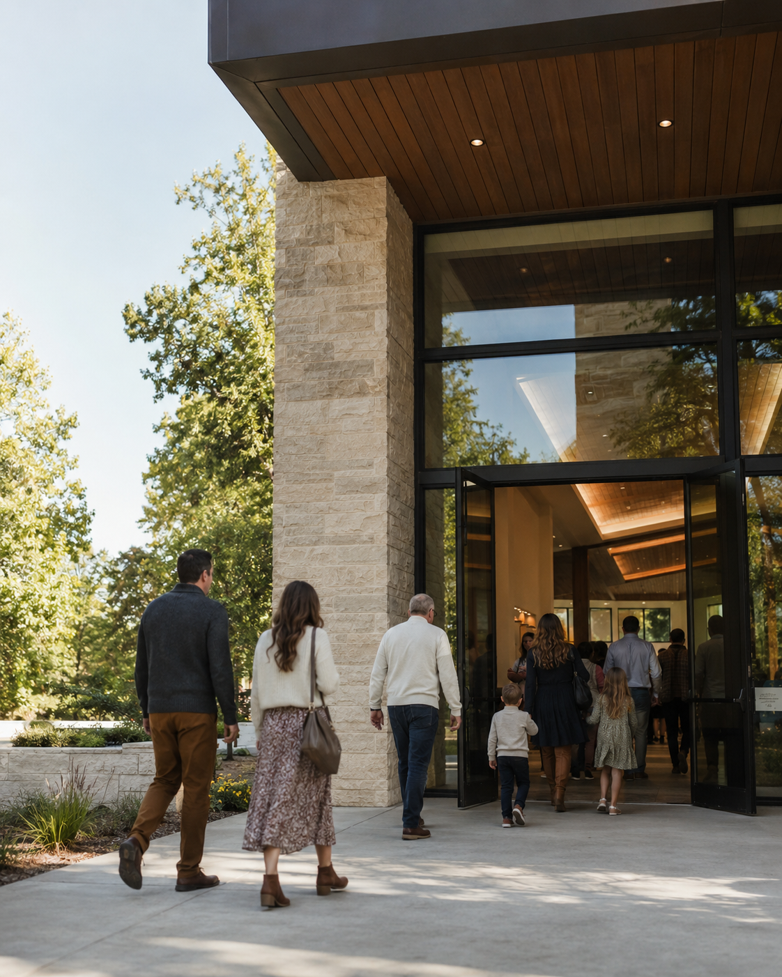 People entering a modern church building with glass doors and stone exterior on a sunny day.