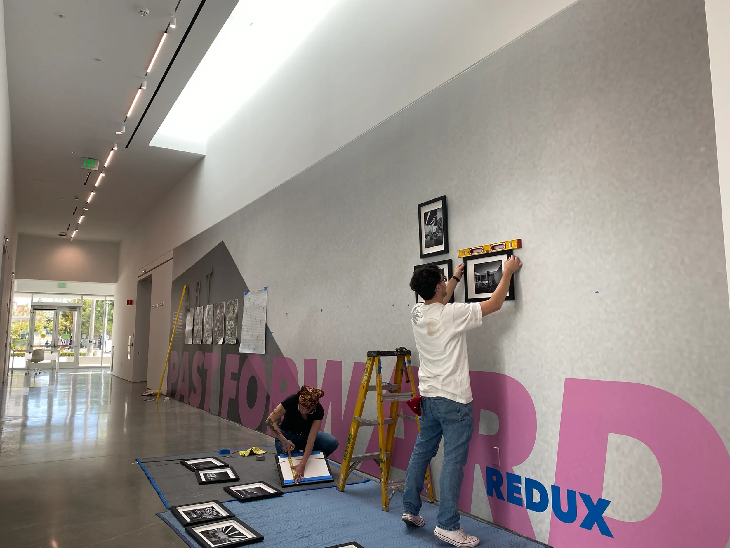 People installing framed photographs on a wall in a building corridor.