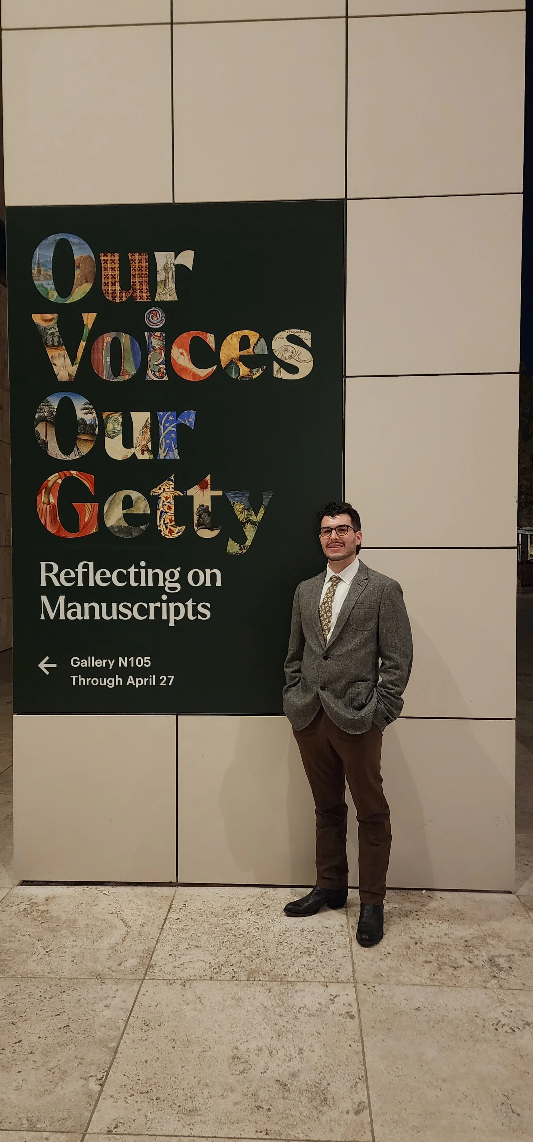A man standing in front of a sign that reads 'Our Voices Our Gritty. Reflecting on Manuscripts' and provides directions to Gallery N105 through April 27. The man is wearing a gray blazer, white shirt, patterned tie, brown pants, and black shoes, with his hands in his pockets.