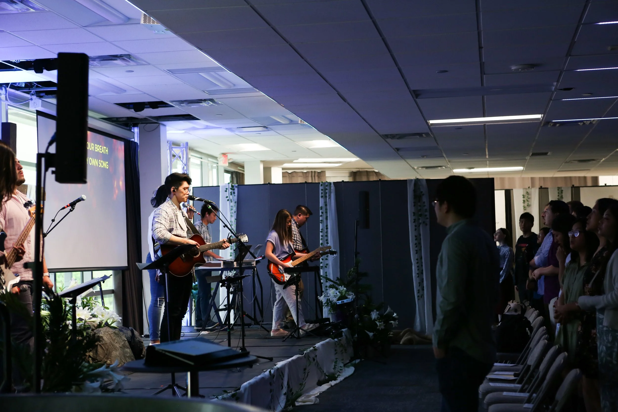 A group of musicians performing on stage during a concert with an audience watching. The stage has microphones, guitars, and a screen displaying song lyrics. The audience is standing and appears to be engaged in the performance.