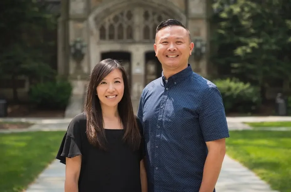 A smiling Asian man and woman standing outdoors on a sidewalk in front of a brick building with arched windows and greenery.