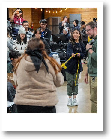 Group of people gathered indoors, with some holding a yellow rope, in a room with wooden walls and string lights.