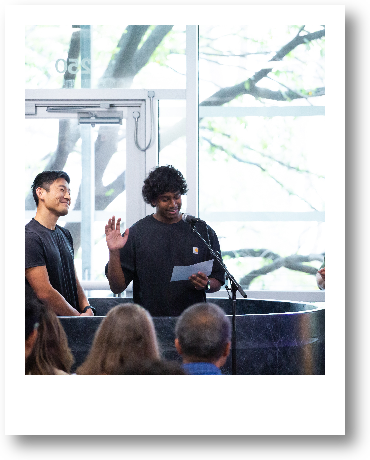 A person with curly hair is giving a speech or presentation at a podium in front of an audience, with a large window and tree visible in the background.