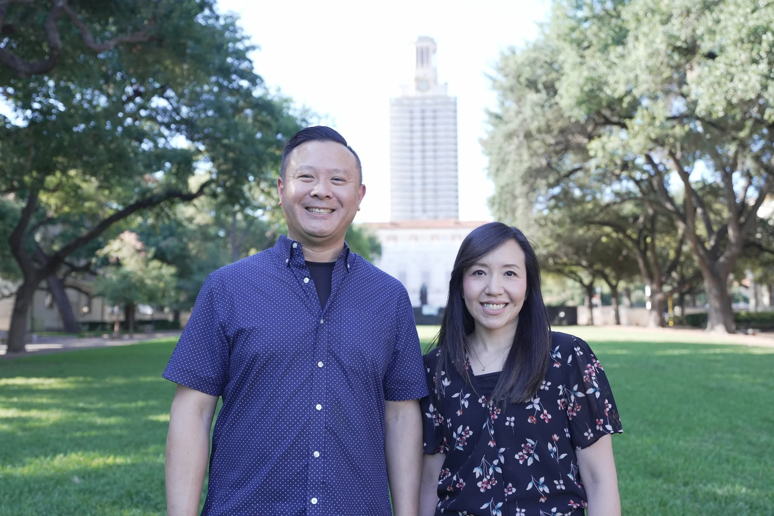 Two smiling people, a man and a woman, standing outdoors in a park with trees and a tall building in the background.