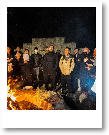 Group of people gathering around a campfire outdoors at night, some wearing masks, with a stone structure in the background and a starry sky above.