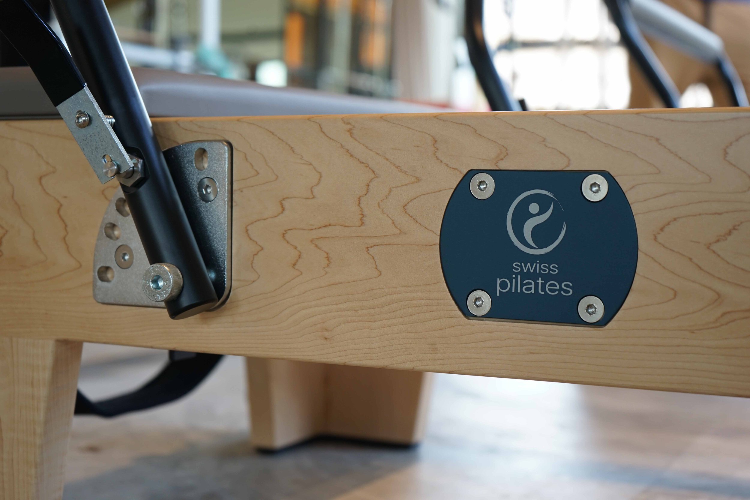 Close-up of a Pilates reformer machine with a wooden frame, metal adjustment components, and a Swiss Pilates logo on the side. The machine is situated in a well-lit studio with a blurred background.