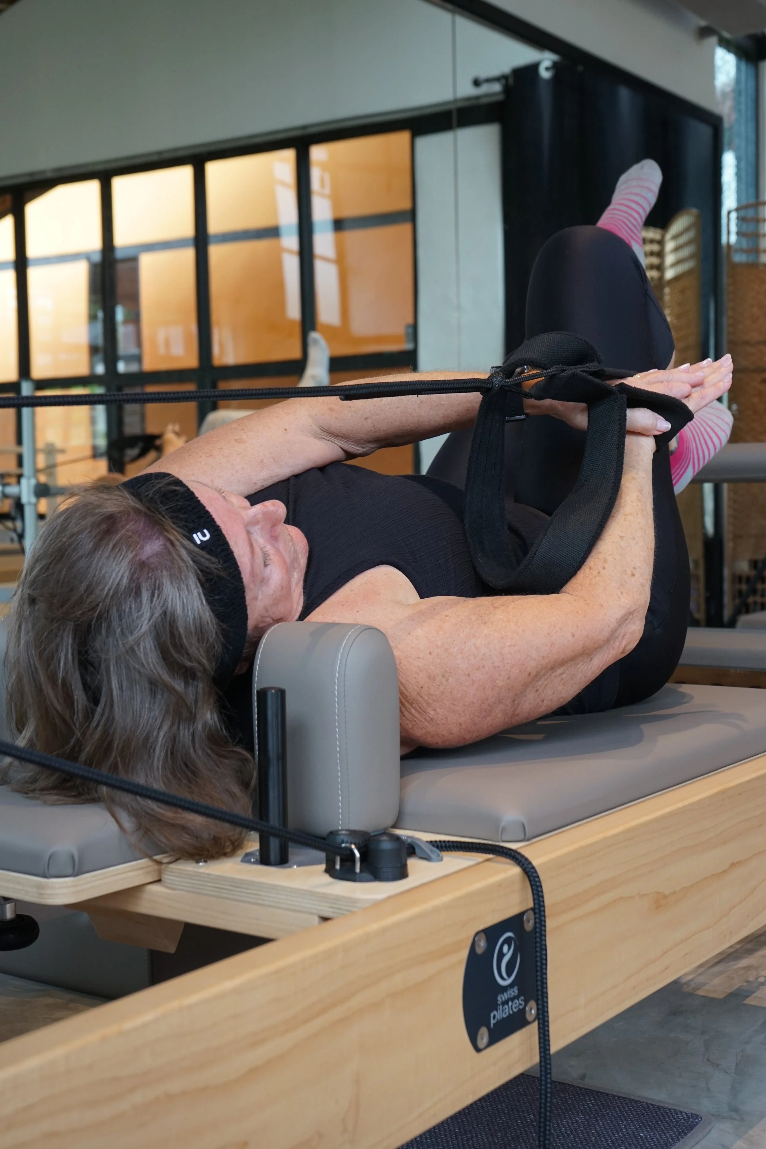 Older woman lying on a Pilates reformer machine during a Pilates class, with her arms extended in front of her and wearing pink and white striped socks.