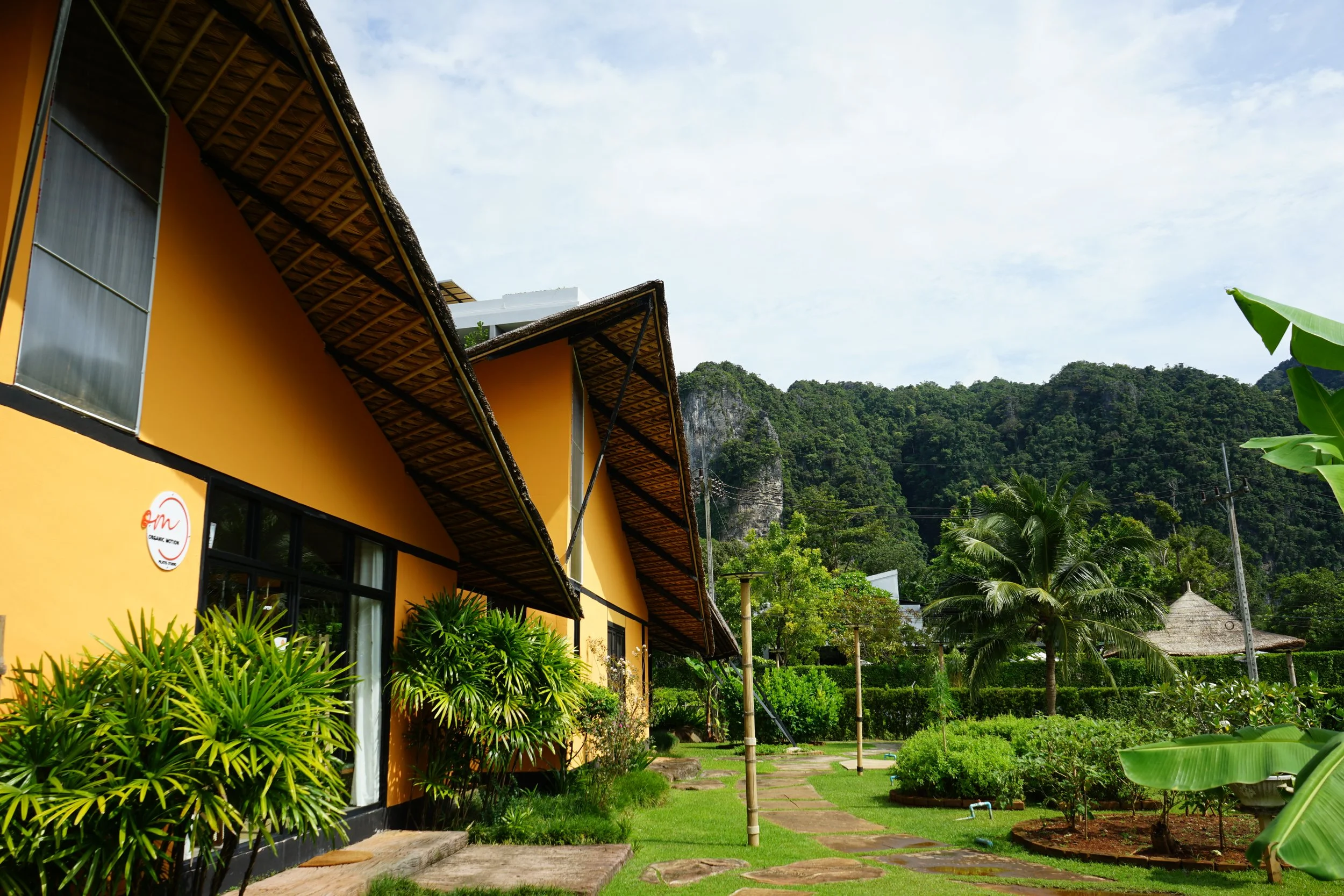 Yellow building with thatched roofs and a lush garden with tropical plants and trees, set against a backdrop of green hills and a partly cloudy sky in Thailand