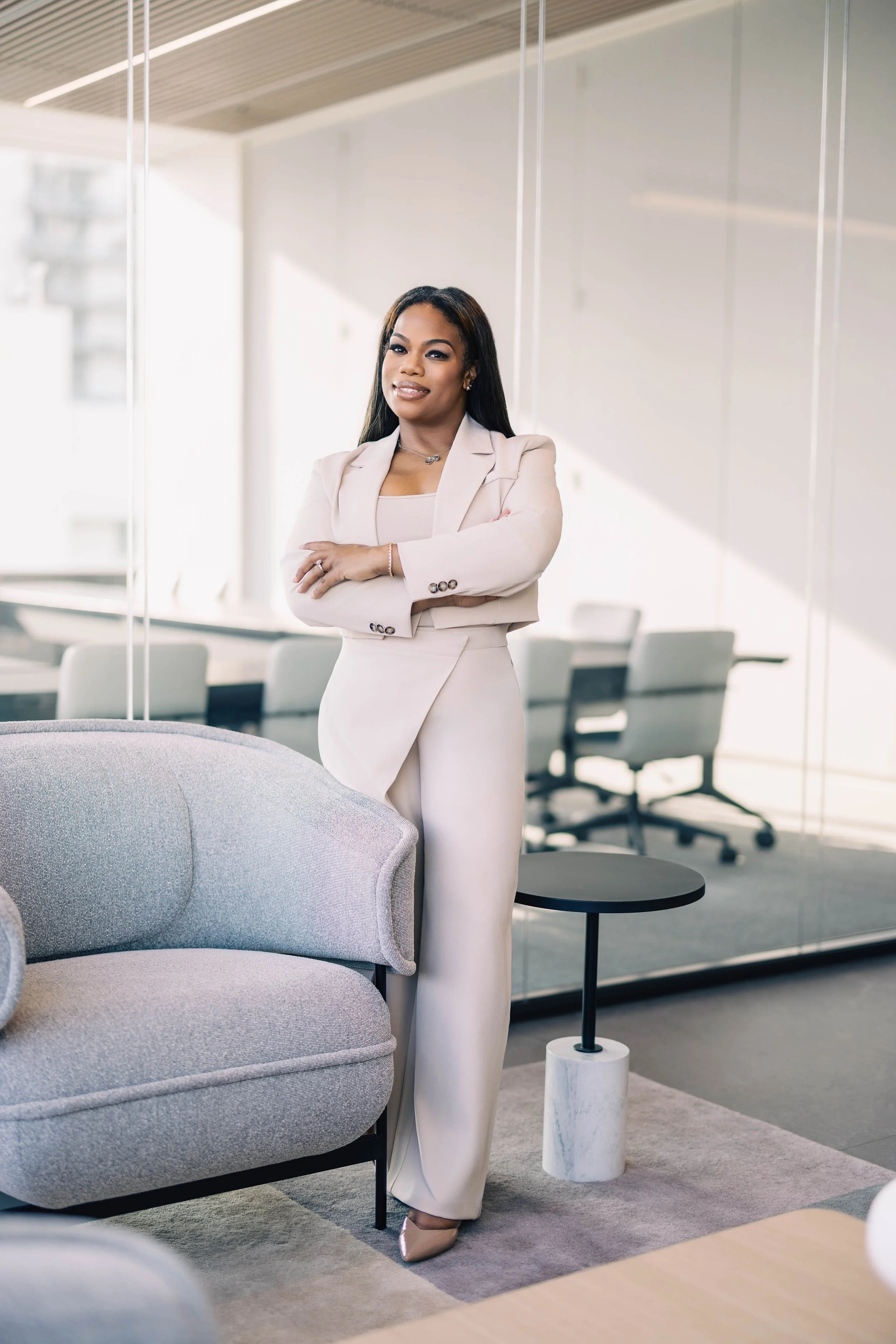 A professional woman standing in an office conference room with crossed arms, wearing a cream-colored suit, beige high heels, and a necklace, near a gray armchair and side table.