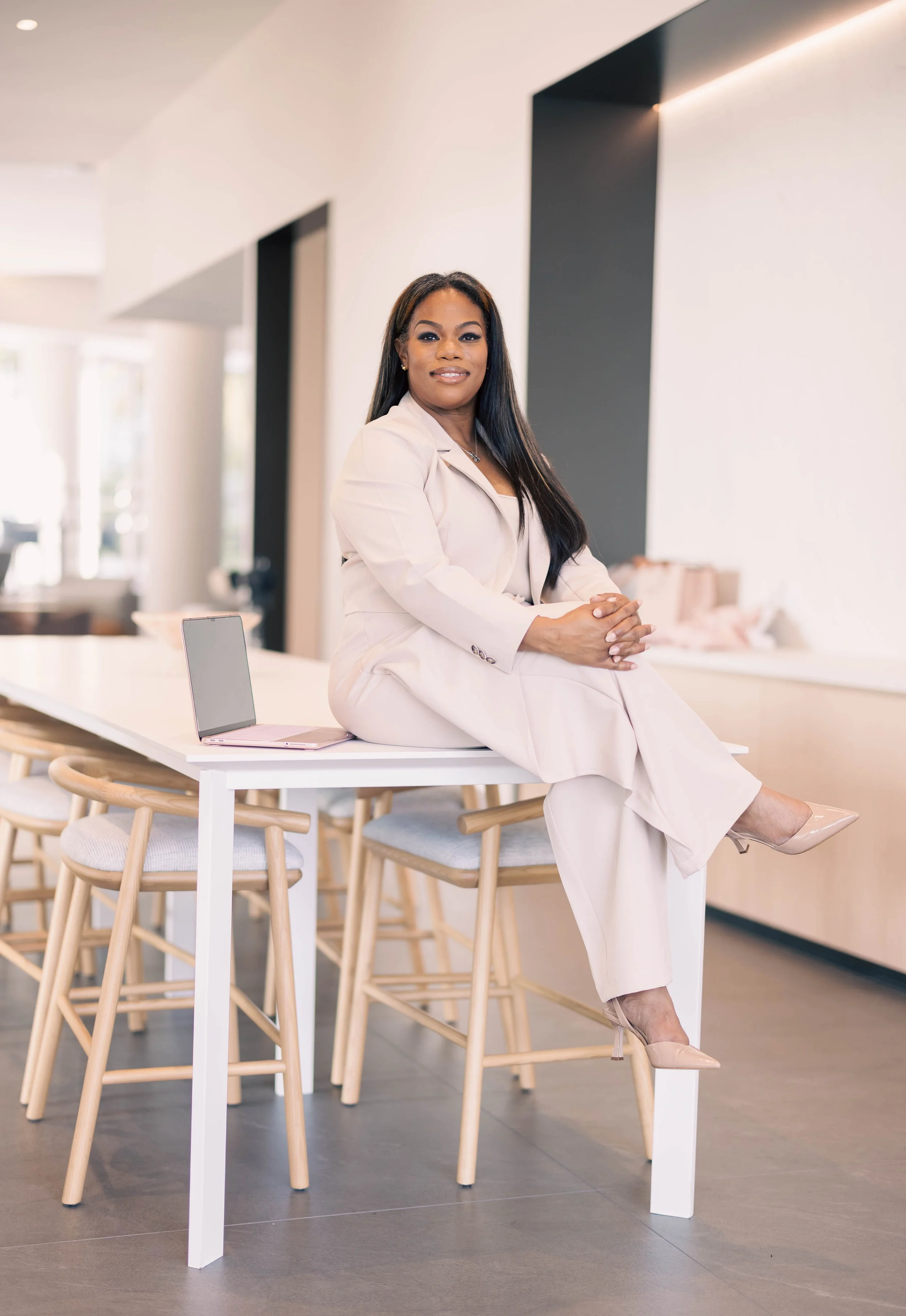 A confident woman with long dark hair, dressed in a light-colored suit, sits on a high table in a modern, minimalist space with natural lighting, wooden chairs, and a laptop nearby.