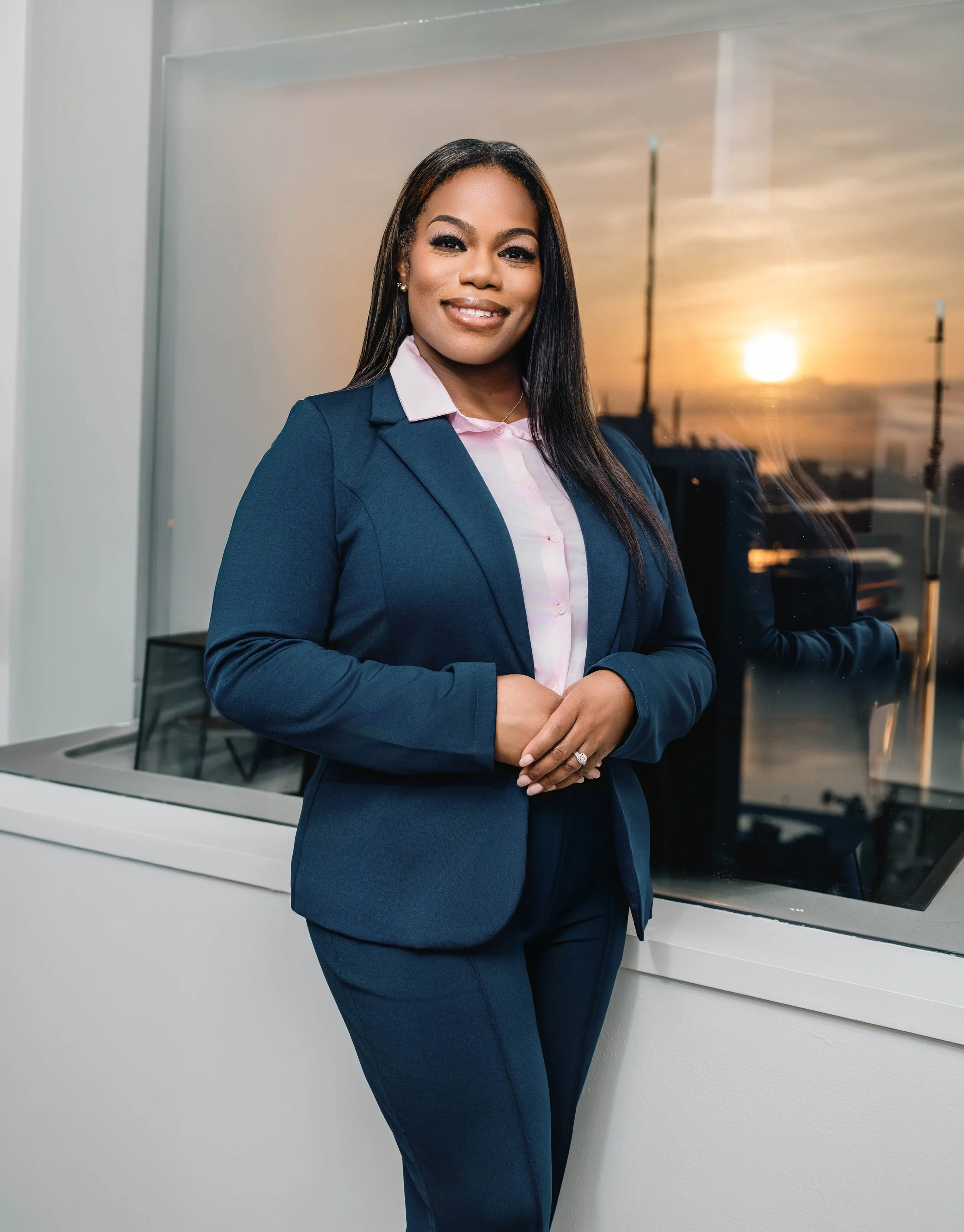 A professional woman in a navy blazer and pink shirt standing in front of a window with a sunset view. She has long straight hair, is smiling, and is wearing rings and earrings.