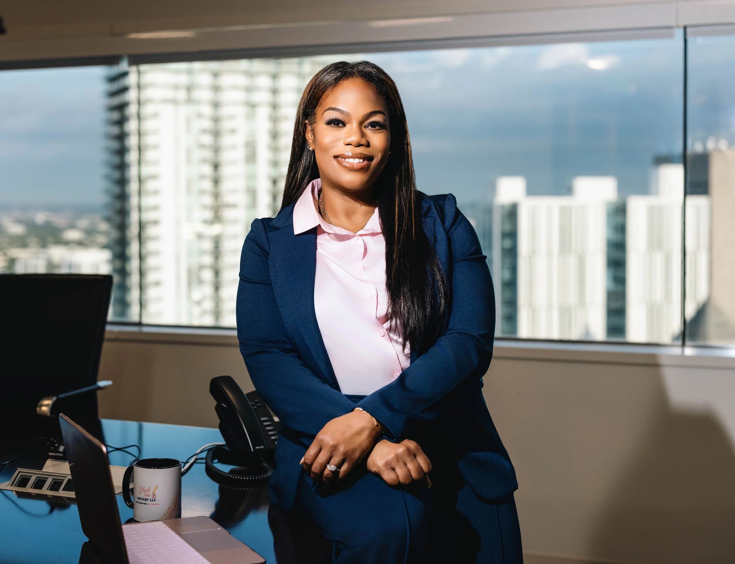 A professional woman in a navy blazer and pink blouse sitting at a conference table with a cityscape view through large windows behind her.