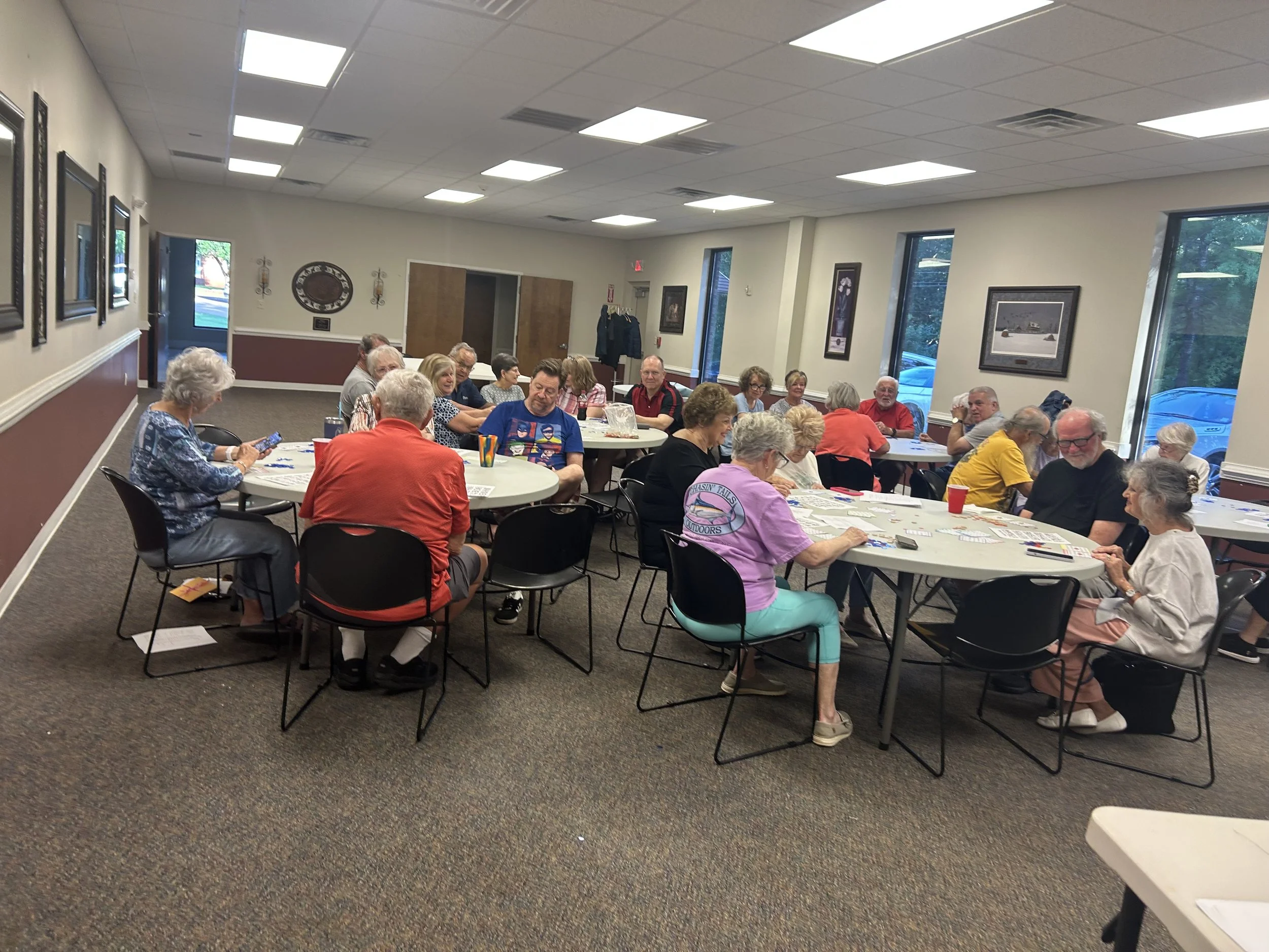 A group of older adults sitting around tables in a community center or meeting room, engaged in an activity or discussion, with some using smartphones and papers spread out on the tables.