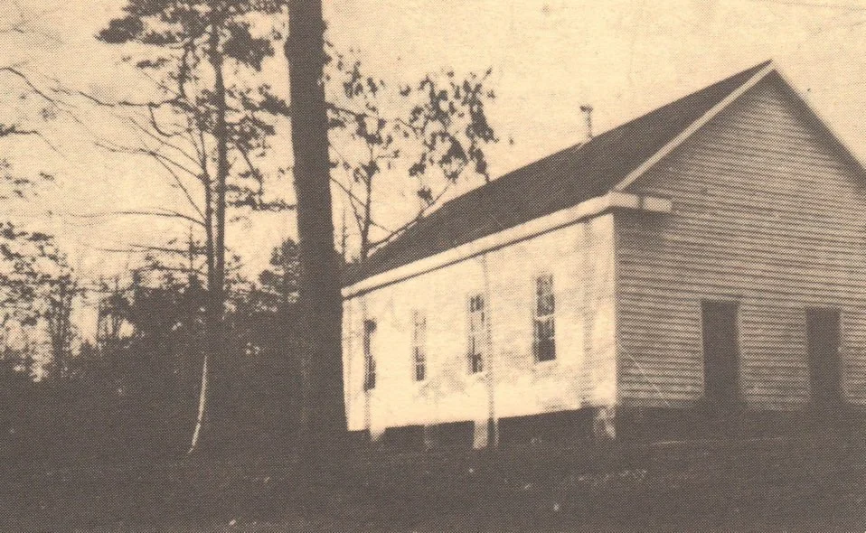 A black and white photo of an old, large wooden house with several windows, surrounded by leafless trees, in a rural setting.