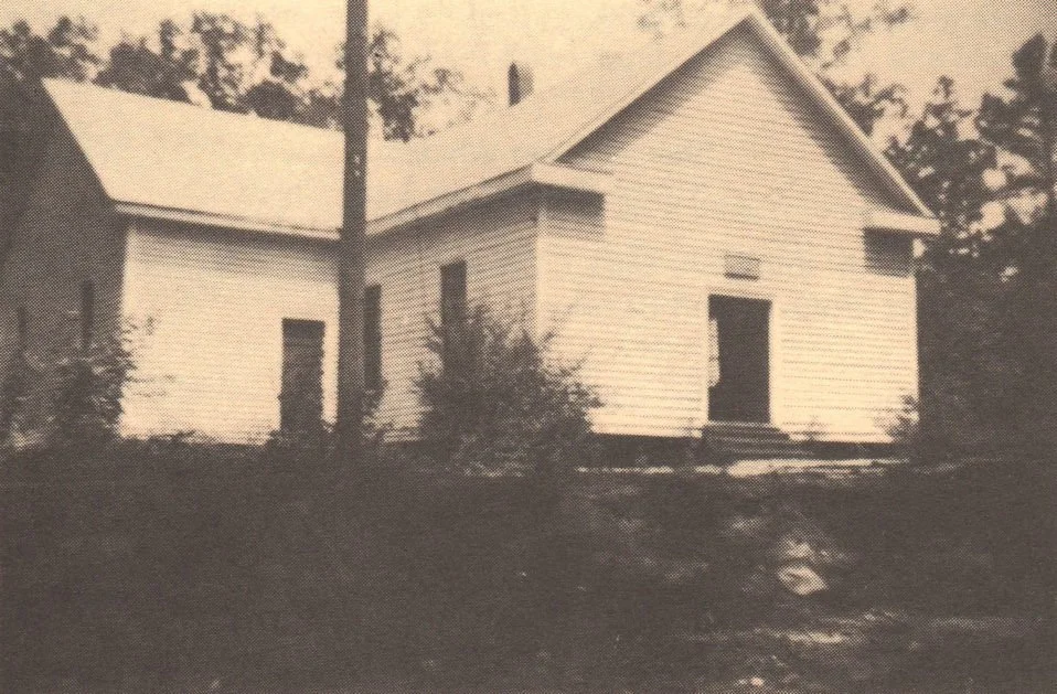 Black and white photo of an old, empty wooden house with a gabled roof, boarded up or missing windows, and bushes in the front yard.