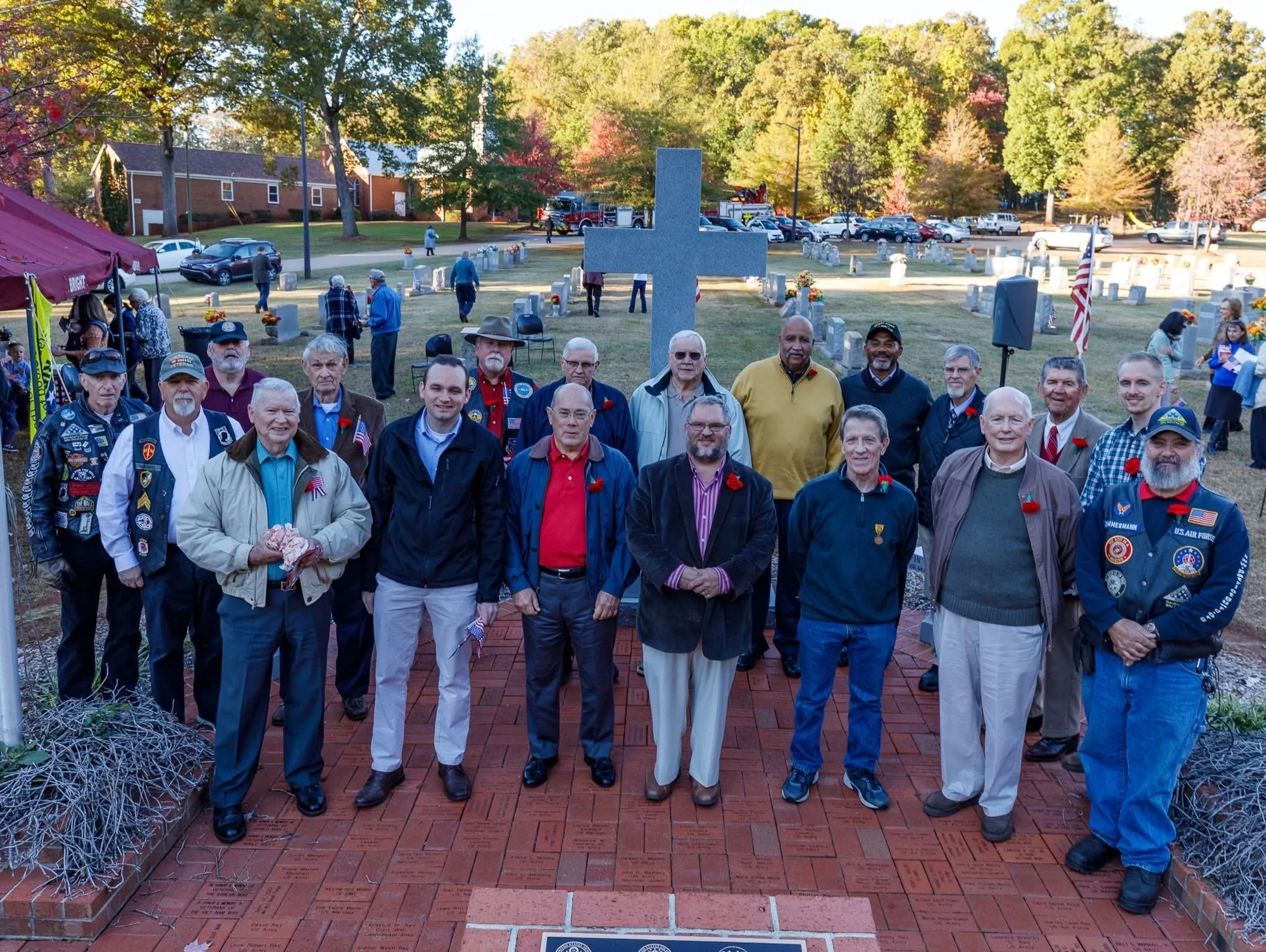 A group of men wearing badges and jackets, standing in front of a memorial with a large granite cross, surrounded by engraved bricks and trees with autumn foliage, during a memorial ceremony in a park.
