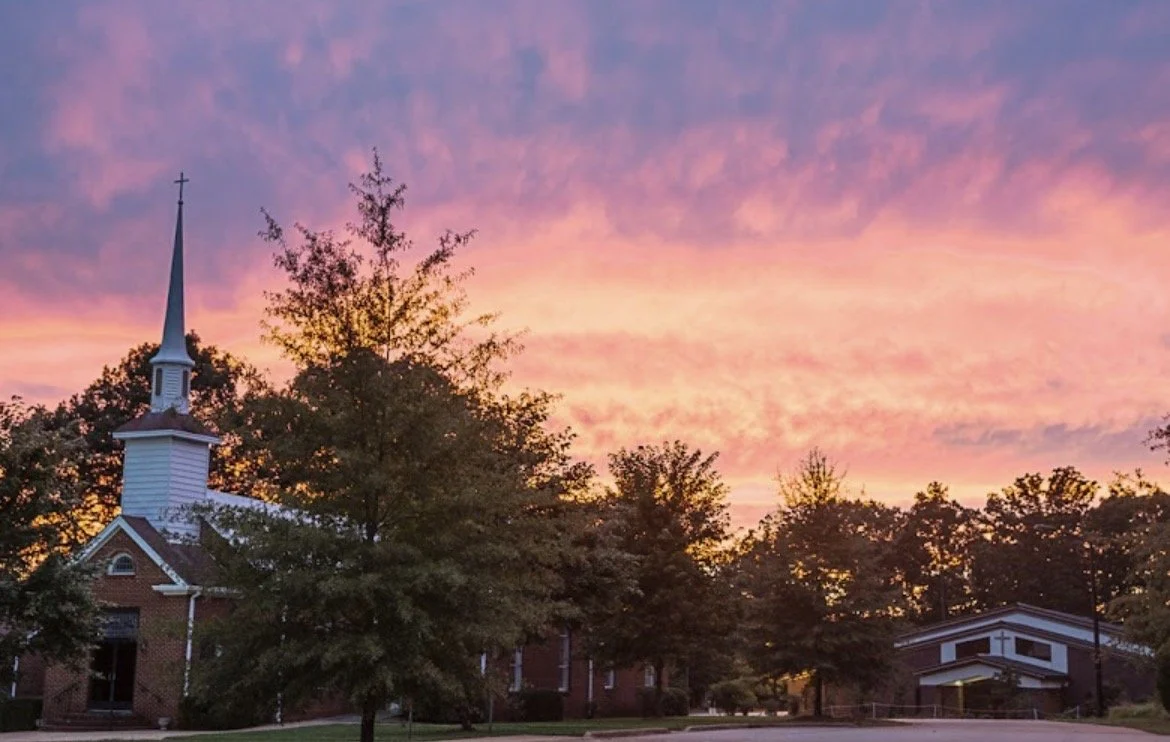 A church with a tall steeple and cross, surrounded by trees, under a colorful sunset sky with pink, purple, and orange clouds.